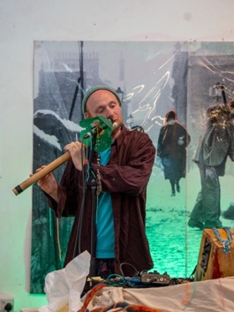 A musician playing a bamboo flute into a microphone in front of a large projected image