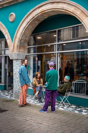 People sitting and standing outside the teal arched shopfront on Newmarket Street, Ayr