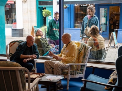 People sitting in armchairs in a shop window talking, with the street visible behind them