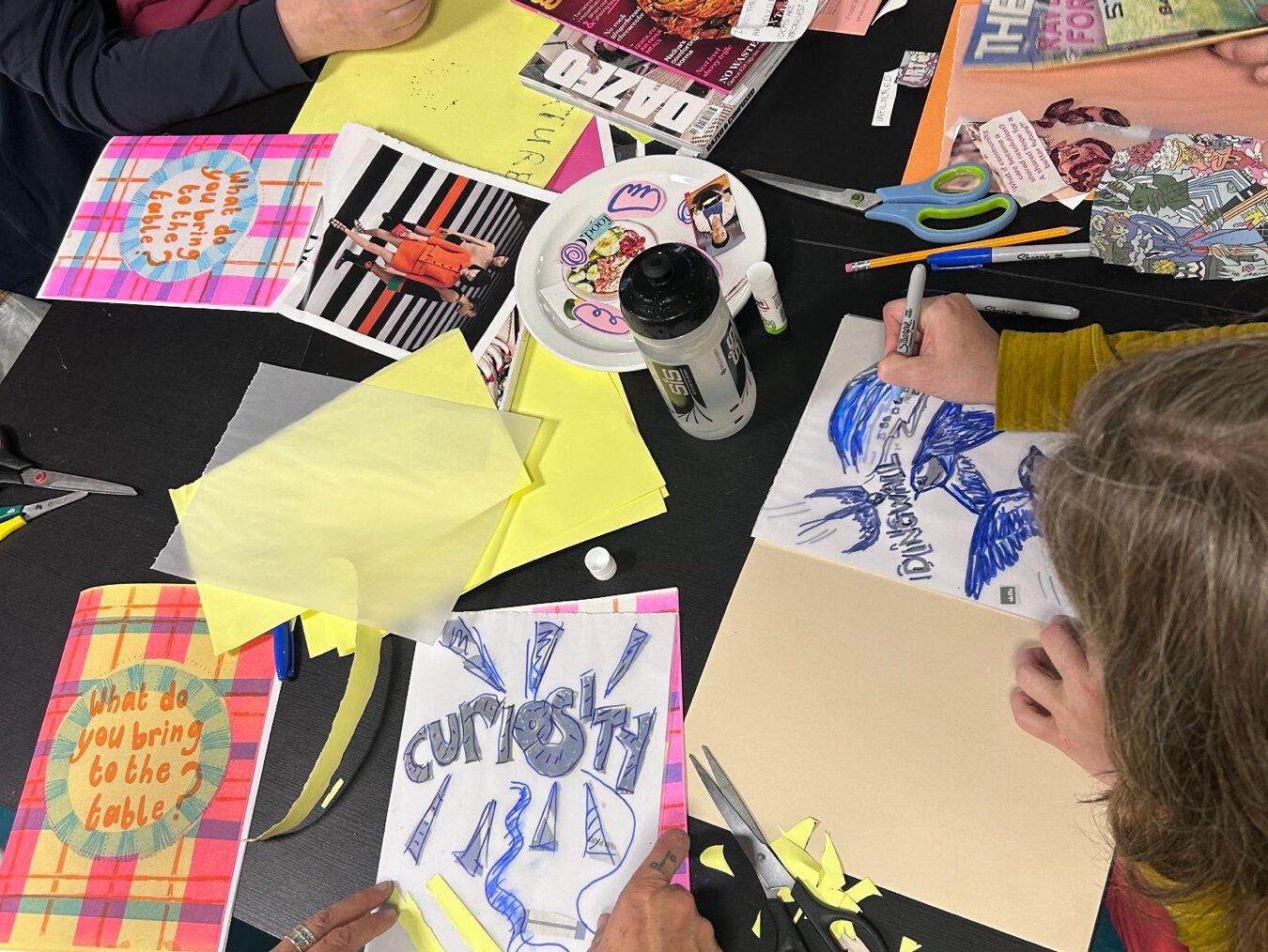 An overhead view of hands cutting and drawing on paper across a table covered in magazines, scissors and zine pages