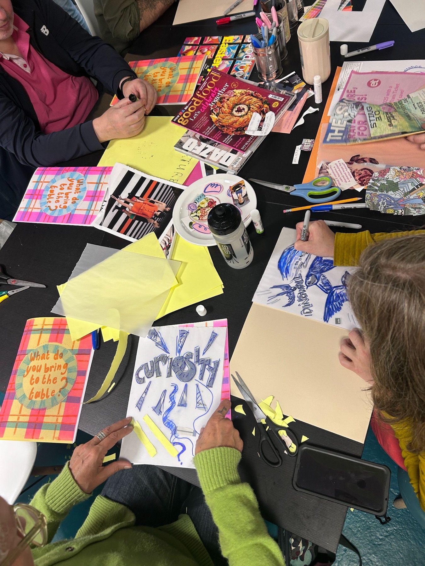 An overhead view of hands cutting and drawing on paper across a table covered in magazines, scissors and zine pages