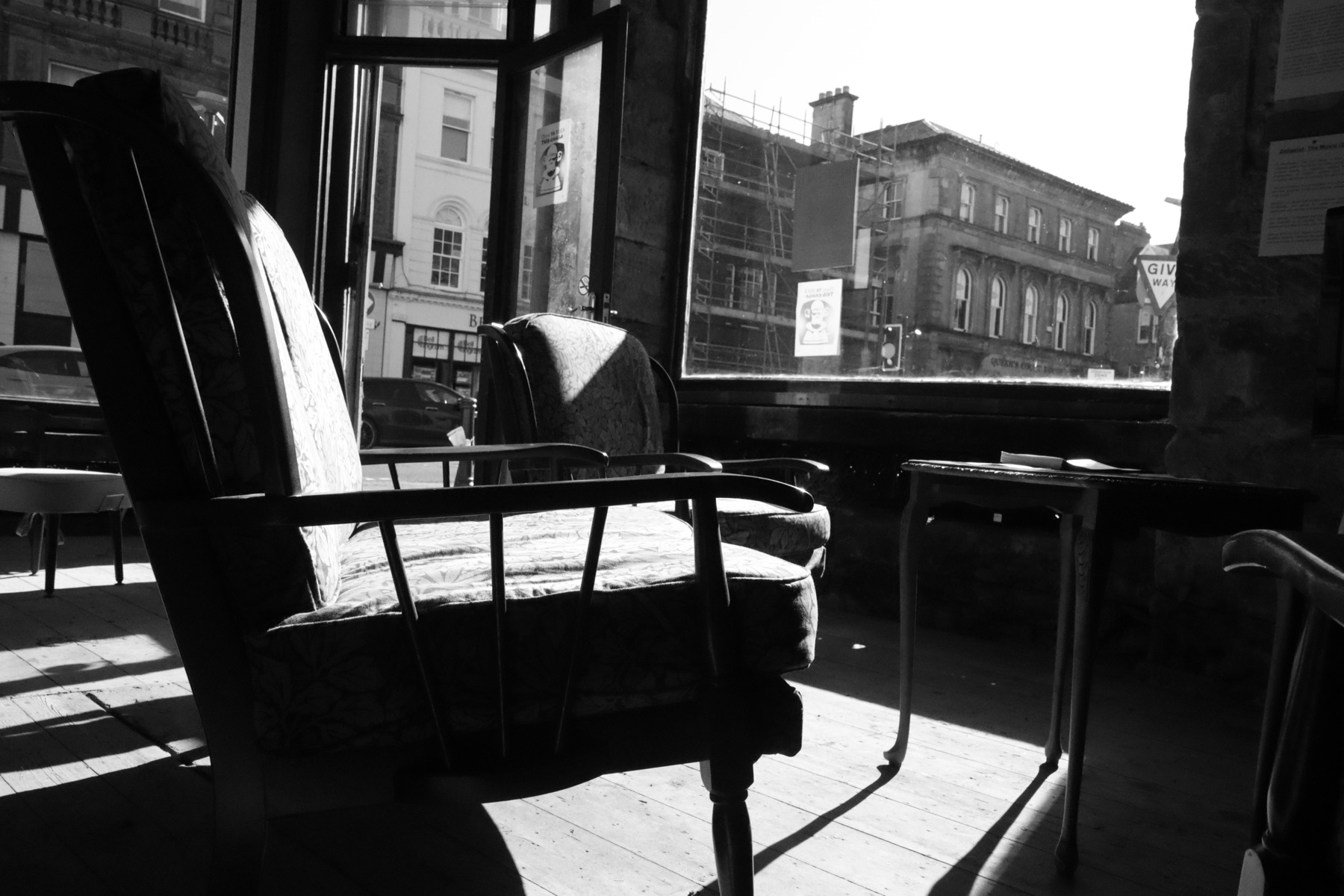 A black and white photograph of armchairs and tables in a bare, sunlit shopfront space looking out onto the street