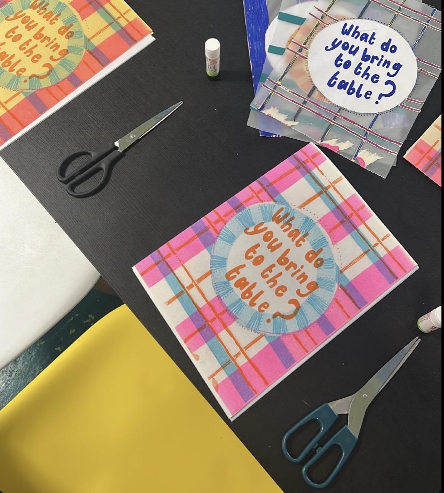 Two-colour Riso printed zine covers reading What do you bring to the table? laid out on a black table with scissors and glue